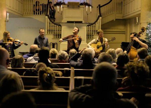 Five musicians at the New Room, with the audience in front of them, sitting in pews. The photo is taken from the audience point of view.