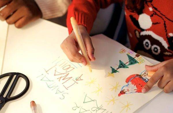 Overhead shot of a craft table with children's hands making Christmas drawing. There's a peek at a Christmas jumper.